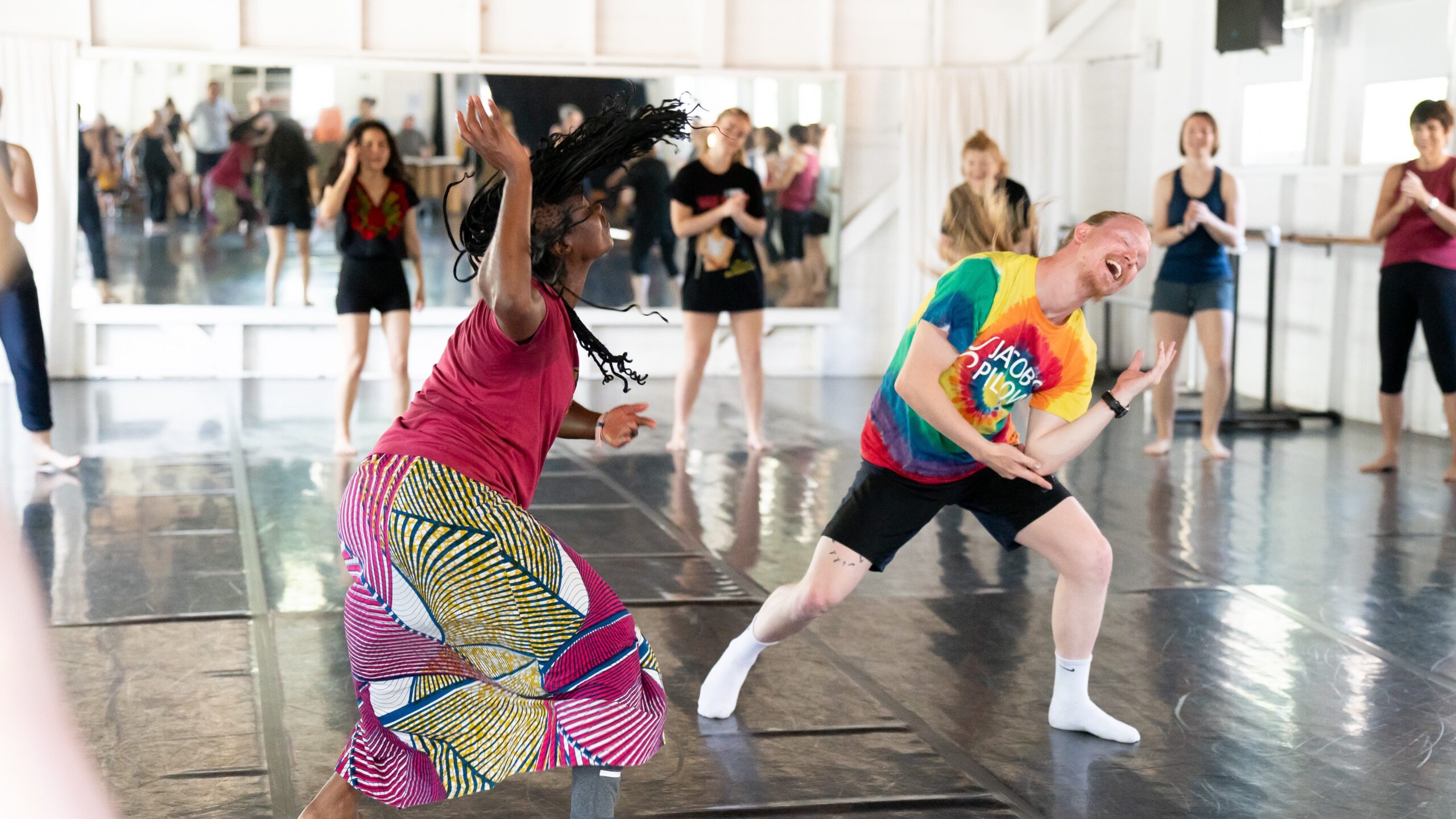 2 dancers appear mid-movement in a West African dance class. The dancer on the left is a middle-aged Black woman wearing a pink t-shirt and patterned skirt. The dancer on the right is red-haired dancer in their early twenties wearing a tie-dyed t-shirt and black shorts.