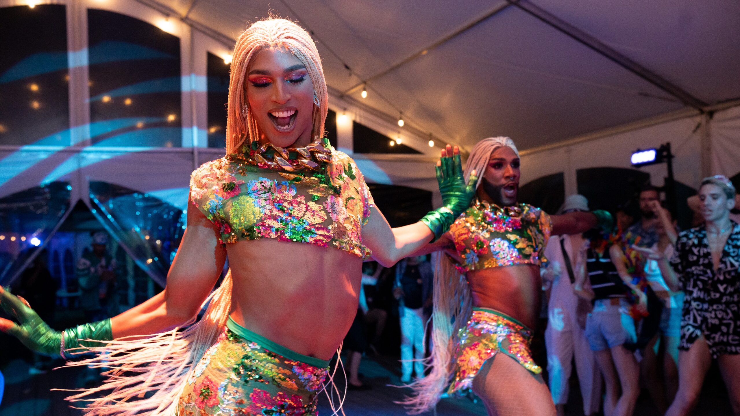 The Dragon Sisters, a drag duo, appear in the middle of their dance at the Jacob's Pillow Pride Party. They are in a tent, and wearing glittery, multi-colored shirts and skirts, as well as long, blonde braids.