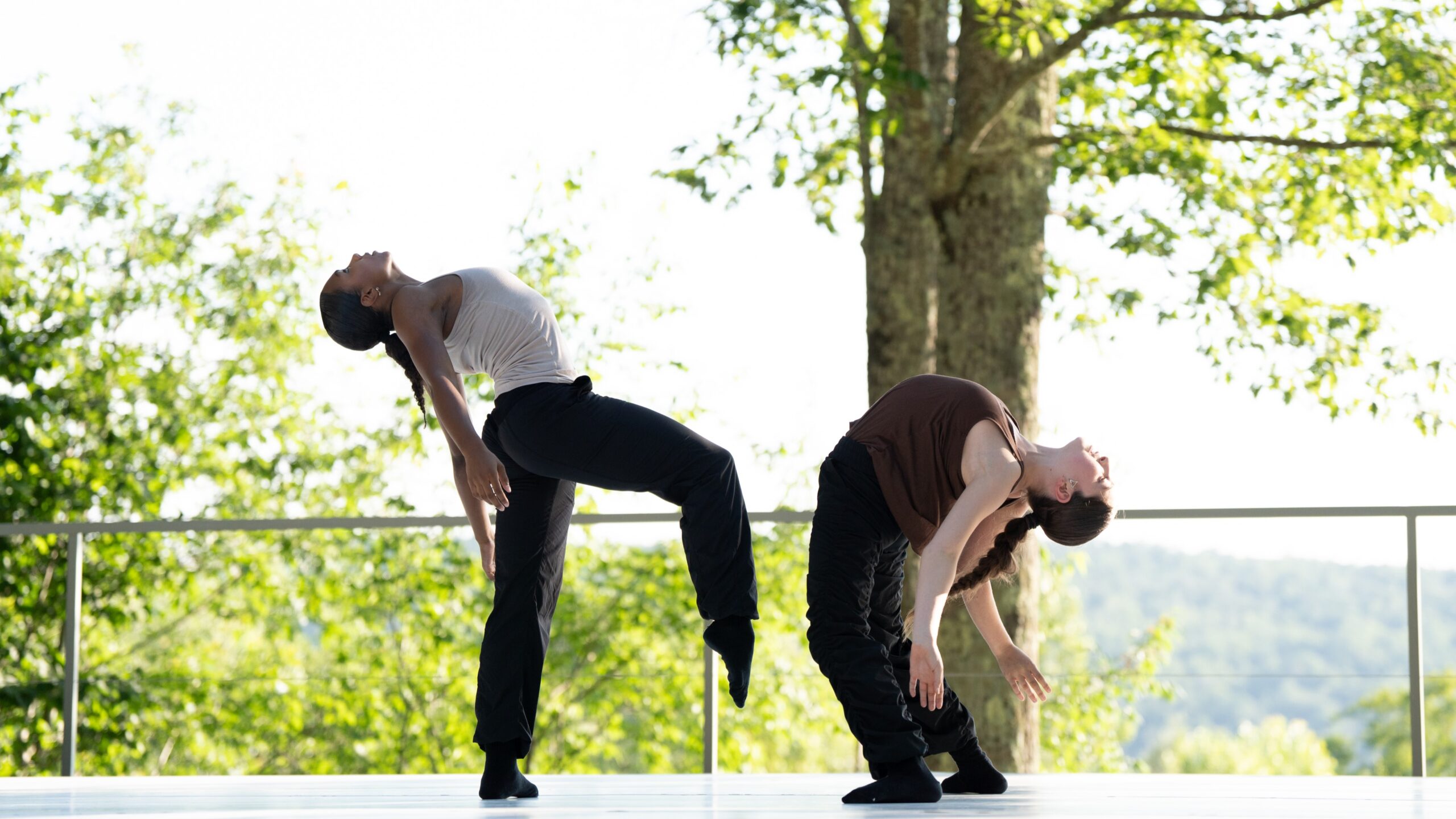 2 female dancers in their late teens or early twenties stand on an outdoor stage. They are wearing tank tops and black pants, and arching their backs to stare at the sky. The dancer on the left is raising her leg slightly off the ground.