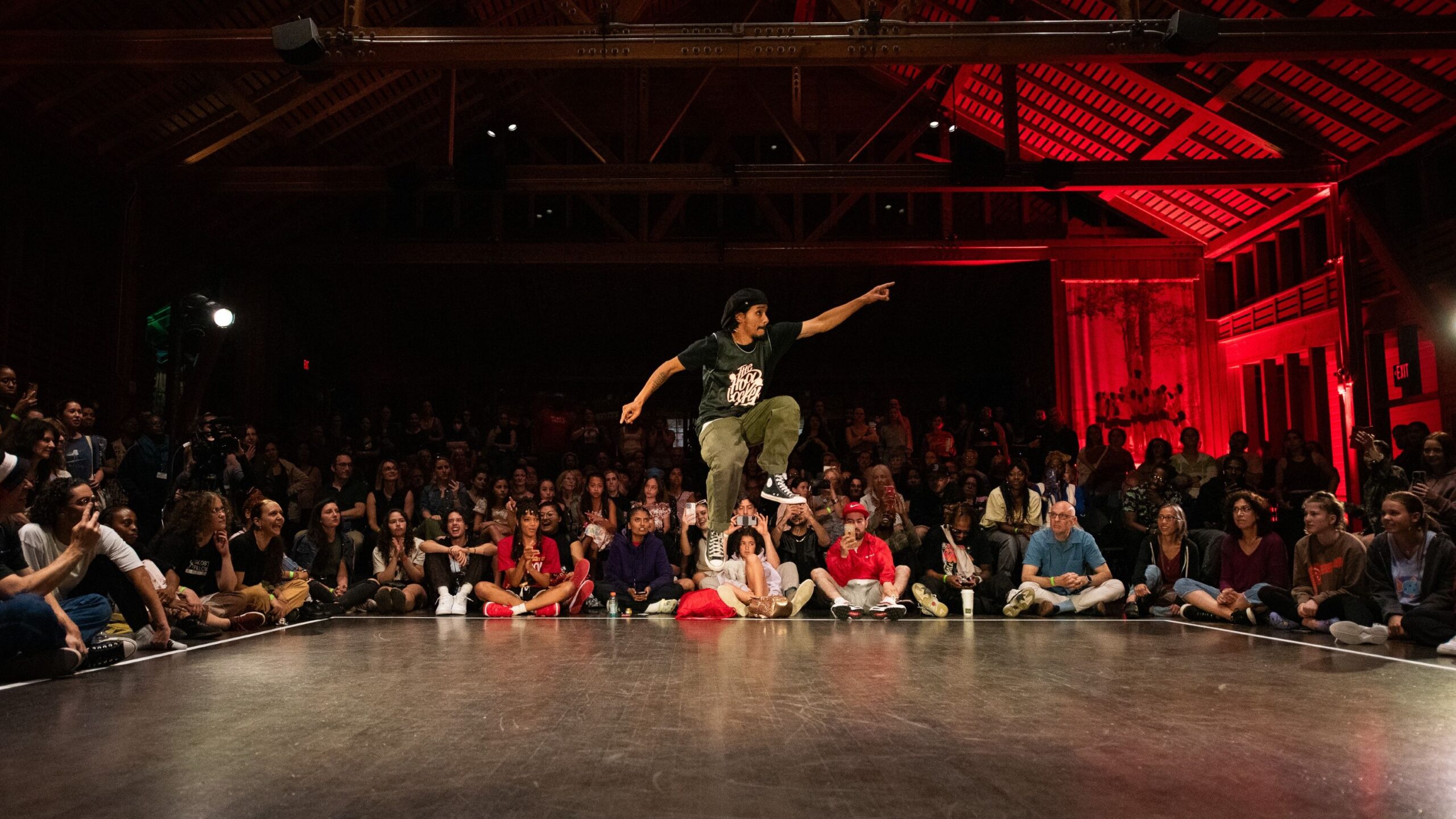 A hip hop dancer wearing a black shirt, black cap, and cargo pants jumps in the air and points to one side. A crowd of people watches on.