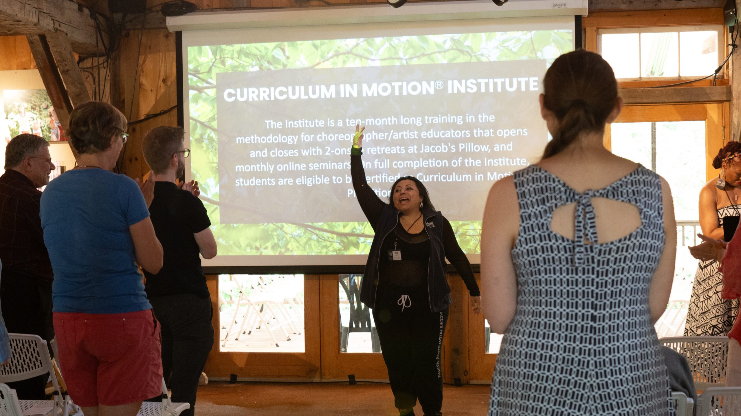 A short woman with dark hair presents to a room of people in front of a screen. The screen is highlighting the Jacob’s Pillow Curriculum in Motion® Institute.