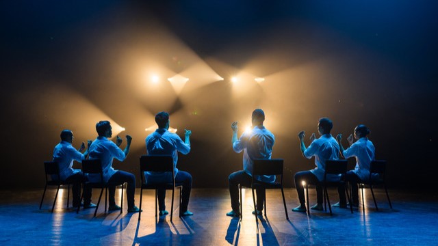 6 dancers appear sitting in chairs with their backs to the camera. They are wearing white, buttoned shirts and sitting with their backs straight and their arms bent in front of their bodies.