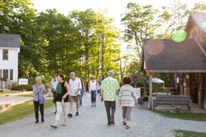 A group of visitors strolls along a sunlit gravel path at Jacob’s Pillow, surrounded by lush green trees and rustic buildings. The warm, golden light and casual conversations among the diverse group of people create a welcoming, relaxed atmosphere typical of a summer day at the festival.