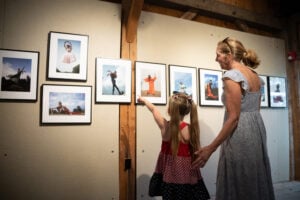 A young girl in a red, white and blue dress excitedly points to a framed photograph on a gallery wall, accompanied by an older woman in a light blue dress, who gently holds her hand. The gallery features vibrant dance portraits, and the moment captures intergenerational curiosity and connection through art.