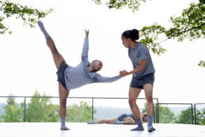 Two dancers dancing on the Henry J. Leir outdoor Stage, wearing light grey shirts, dark gray short, and light gray socks. The dancer on the left has their leg extended and the dancer on the right is supporting them.