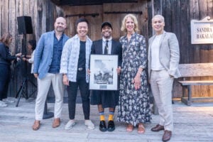 Five people pose together on a wooden deck in front of a rustic wooden building. The person in the center, wearing a black suit jacket, tie, skirt, and a baseball cap, holds a framed black-and-white award or photograph and smiles proudly. The others, dressed in semi-formal attire including blazers, a floral dress, and light-colored suits, stand closely and smile warmly. The mood is celebratory and supportive.